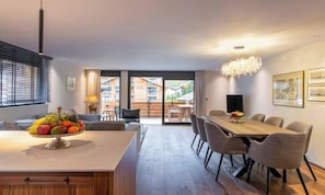 A modern kitchen with a central island, dining area with a long table, and a view of a balcony with outdoor seating. - LUXURY ALPINE APARTMENT WITH SPA, TECHNOGYM & PRIVATE CINEMA (Zermatt)