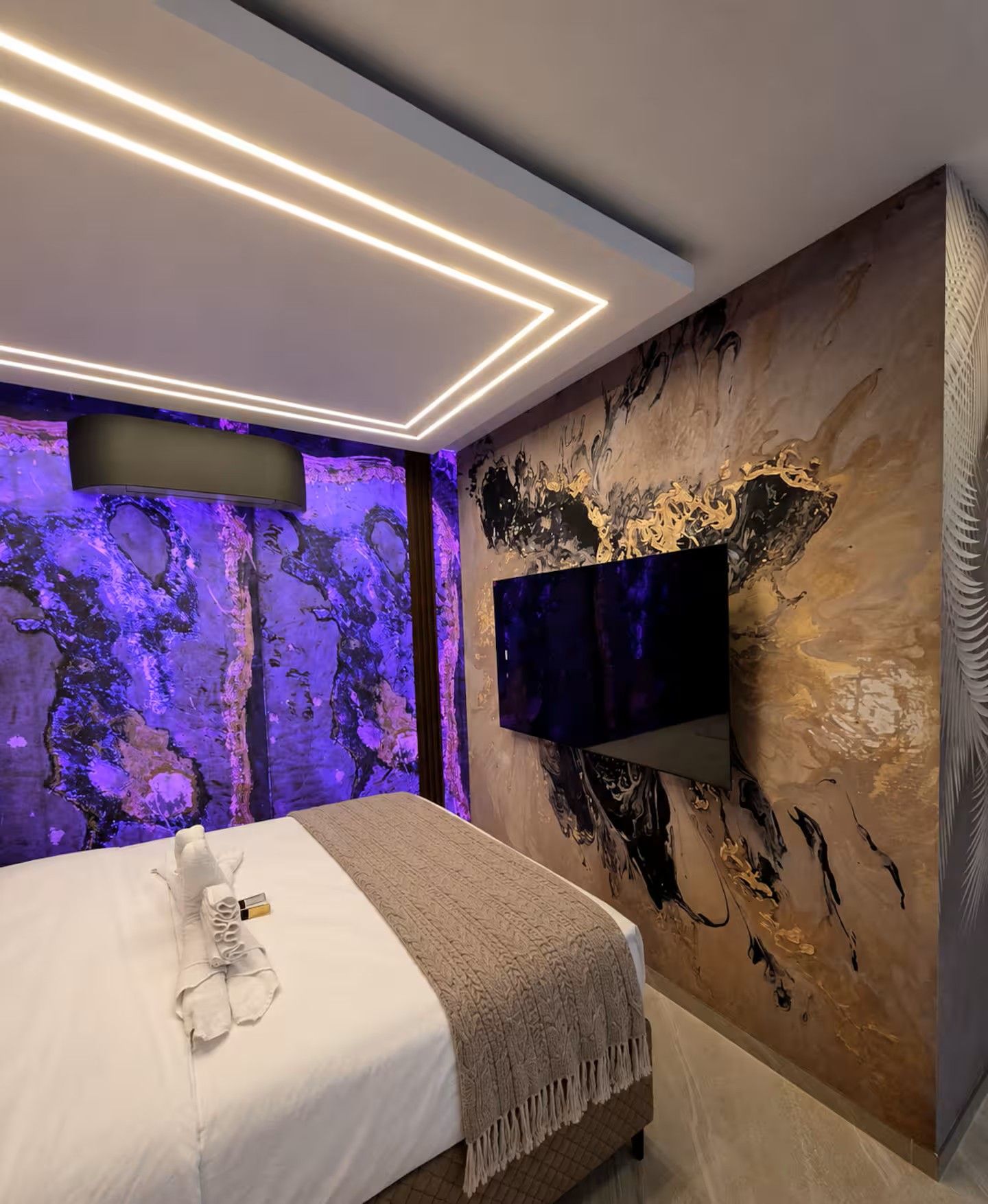 Une chambre à coucher moderne, avec un lit, un téléviseur à écran plat fixé au mur et un éclairage d’ambiance.