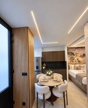 Un intérieur moderne comprenant un coin repas avec une table ronde et des chaises, une cuisine avec des meubles sombres et un salon avec un canapé et des œuvres d’art décoratives au mur.