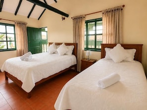 A hotel room with two beds, a wooden headboard, a nightstand, a window with curtains, and a door. - Hotel Altair (Villa de Leyva)