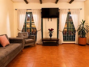 A living room with a sofa, a TV mounted on the wall, a potted plant, and a window with curtains. - Hotel Altair (Villa de Leyva)