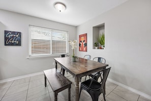 A dining area with a wooden table, benches, and chairs. There are two wall decorations, one with a plant and another with a floral design. - Luxury Escape - Pool, Hot Tub, Arcade, Games, BBQ & Outdoor Lounge! (Plano)