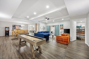 A spacious living room with a pool table, a blue sofa, an orange armchair, and a dining area with wooden chairs. - Luxury Escape - Pool, Hot Tub, Arcade, Games, BBQ & Outdoor Lounge! (Plano)