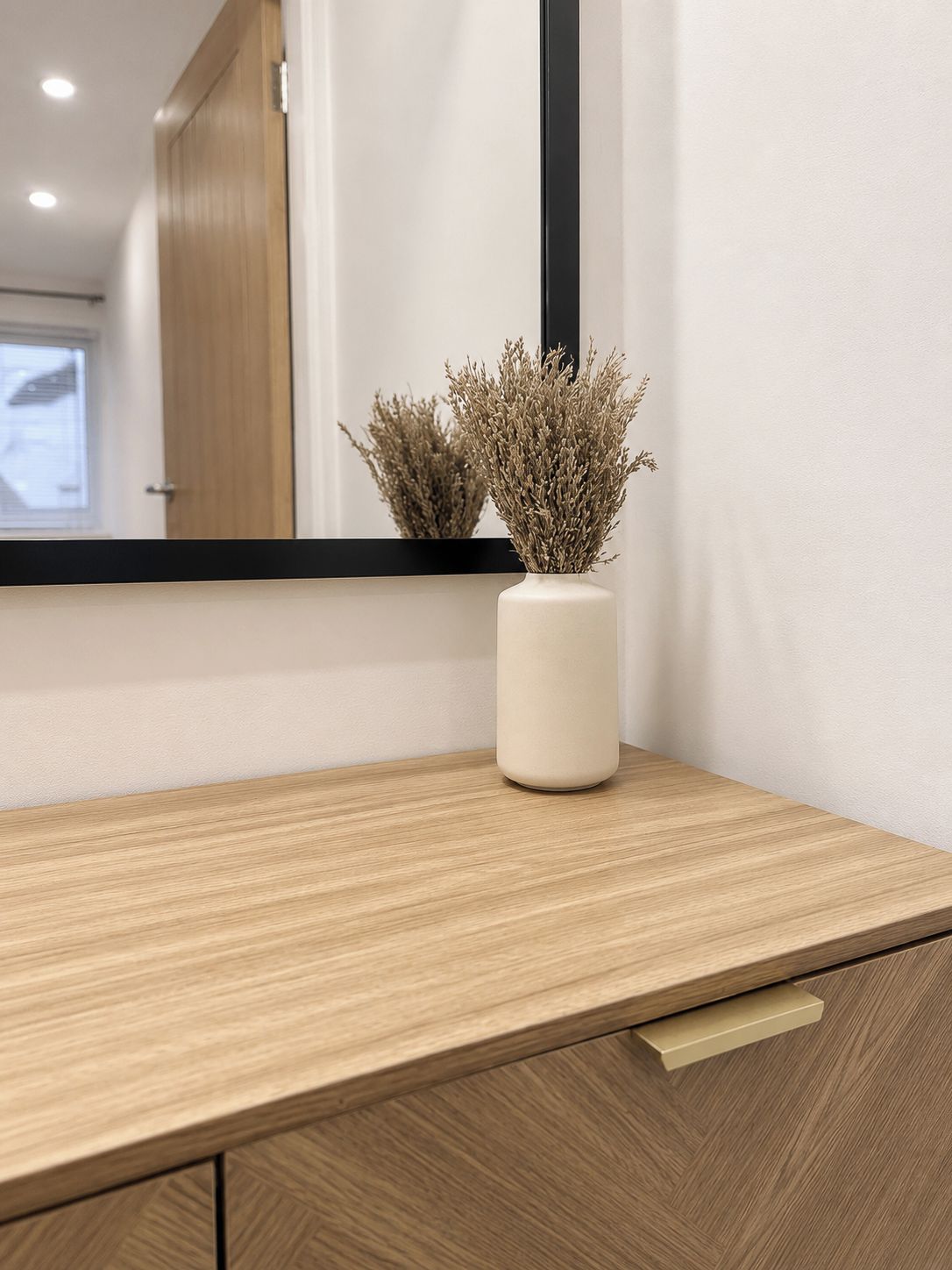Una encimera de madera con un jarrón blanco que contiene plantas secas, un espejo y una puerta al fondo.