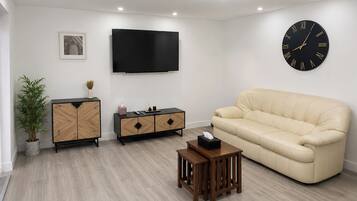 Una sala moderna con una televisión de pantalla plana, un reloj de pared grande, un sofá color beige, un mueble de madera y una pequeña mesa de madera.