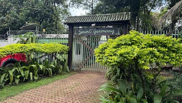 Una entrada protegida por un portón con un letrero que dice 'Saint Germany', rodeada de abundante vegetación y un camino pavimentado.