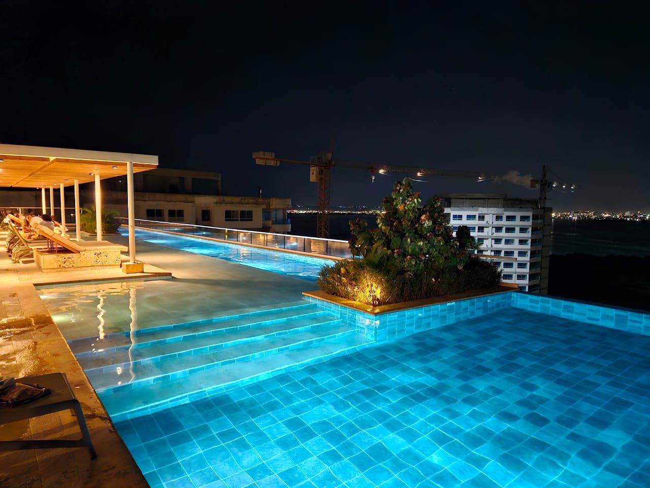 A nighttime swimming pool with steps leading into it, surrounded by lounge chairs and a small garden area.