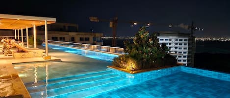 A nighttime swimming pool with steps leading into it, surrounded by lounge chairs and a small garden area.
