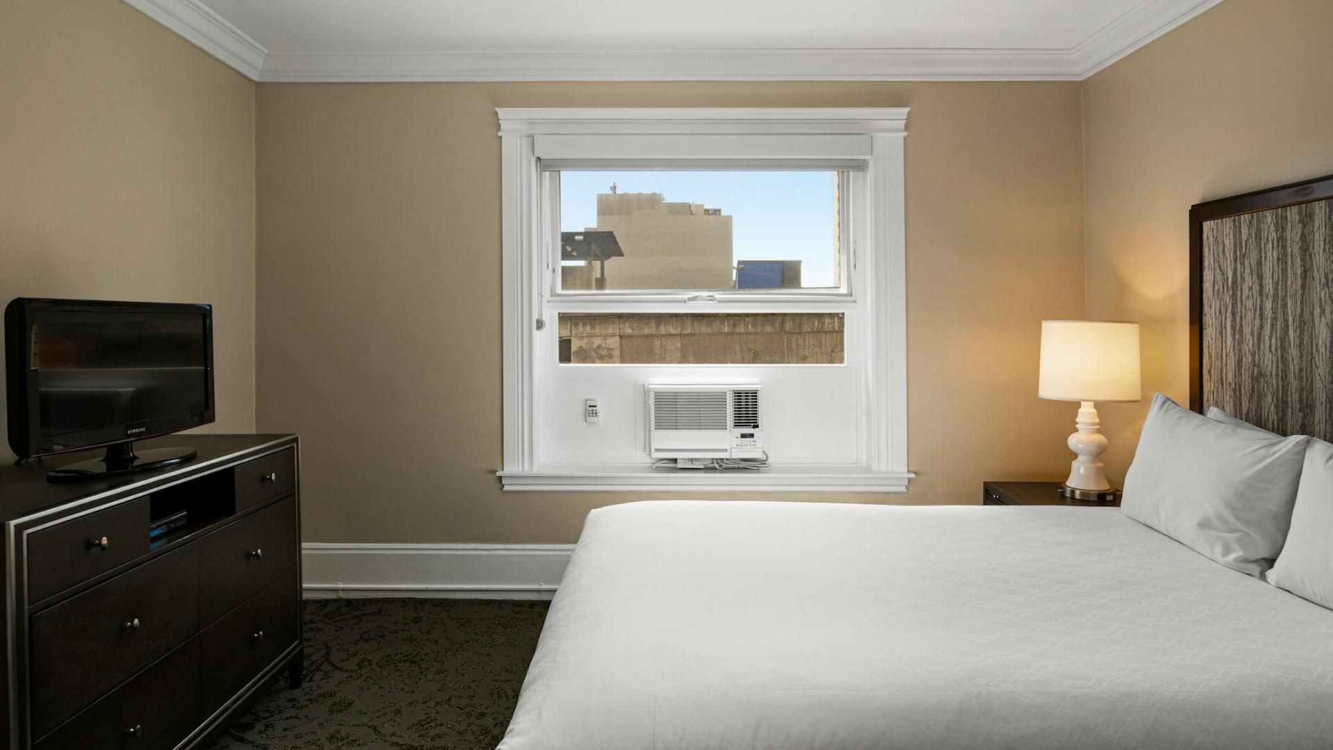 A hotel room with a bed, a dresser, a television, a window with an air conditioner, and a lamp.