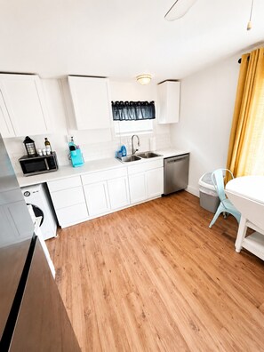 A compact kitchen with white cabinets, a microwave, a sink, and a dishwasher. There's a small table with a chair and a yellow curtain. - Lake Escape Cottage | Minutes to Paw Paw Lake + Wineries (Watervliet)