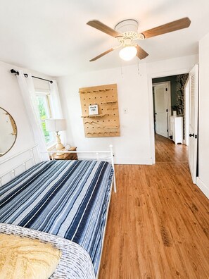 A bedroom with a bed, a ceiling fan, a wall-mounted wooden board, and a door leading to another room. - Lake Escape Cottage | Minutes to Paw Paw Lake + Wineries (Watervliet)