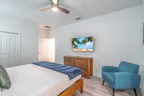 Une chambre à coucher comprenant un lit, une commode, une chaise, un ventilateur de plafond et un téléviseur fixé au mur.