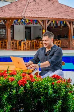 Una persona sentada junto a una piscina con una computadora portátil, rodeada de vegetación y un edificio con tejado de baldosas.