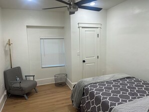 Une chambre à coucher comprenant un lit, une chaise, un ventilateur de plafond, une fenêtre avec des stores et une porte.