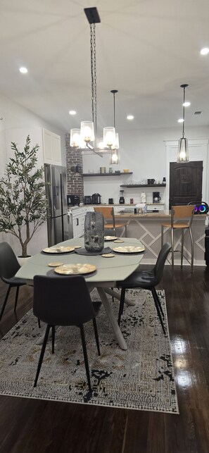 Un comedor moderno con una mesa central, sillas, una lámpara colgante y una cocina al fondo.