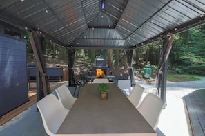 An outdoor covered patio with a long table and chairs, a fire pit, and a grill. - Hidden Dome Retreat in the Trees (Coos Bay)