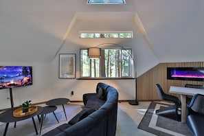 A modern living room with a sofa, a TV, a dining area with chairs, and a window with a view of trees. - Hidden Dome Retreat in the Trees (Coos Bay)