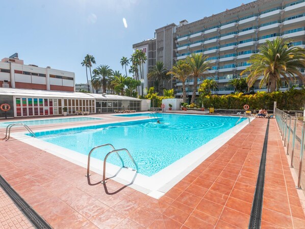 A swimming pool with a tiled deck, surrounded by palm trees and a building with multiple balconies. - Tropical Views - Playa del Inglés (Playa del Inglés)