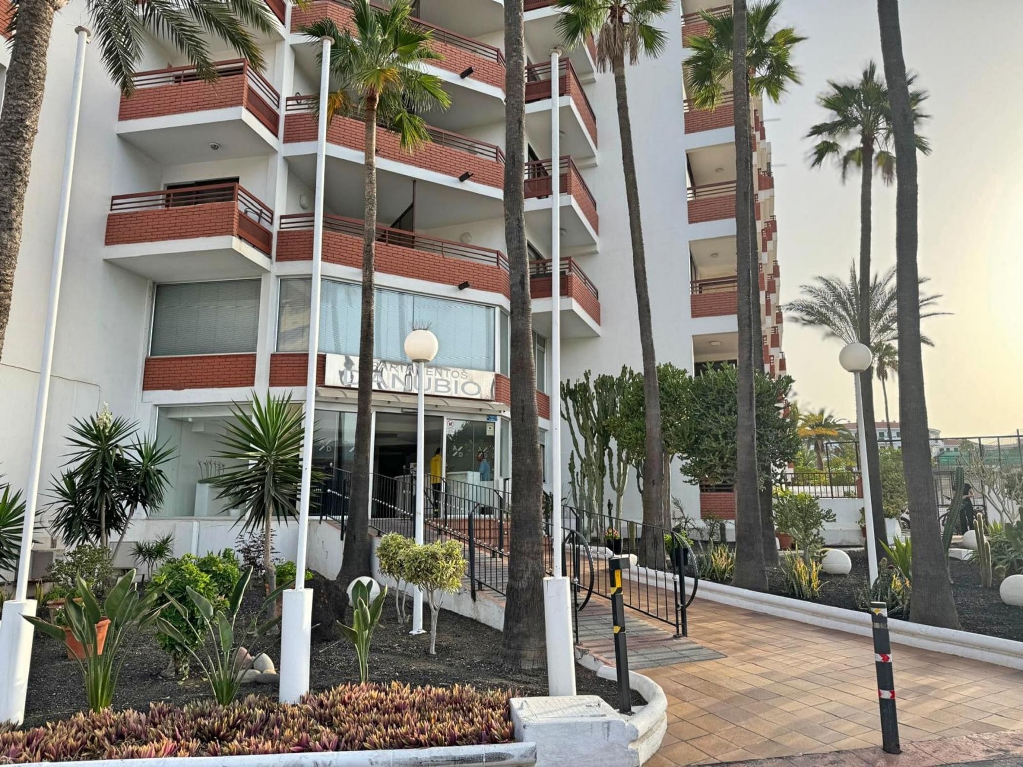 Un edificio de varios pisos con balcones, un letrero que dice "PARQUE DE LOS VIEJOS" y un área ajardinada con palmeras y plantas.