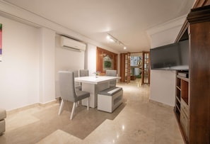 Un comedor moderno con una mesa blanca y sillas grises, un televisor de pantalla plana a la derecha, y un mueble de madera abajo.