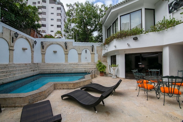 Una piscina con sillas de descanso y una mesa, rodeada por un edificio blanco con ventanas arqueadas y un balcón.