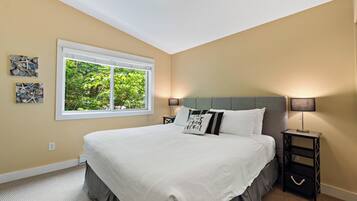A bedroom with a large bed, a window with a view of greenery, and a nightstand with a lamp.