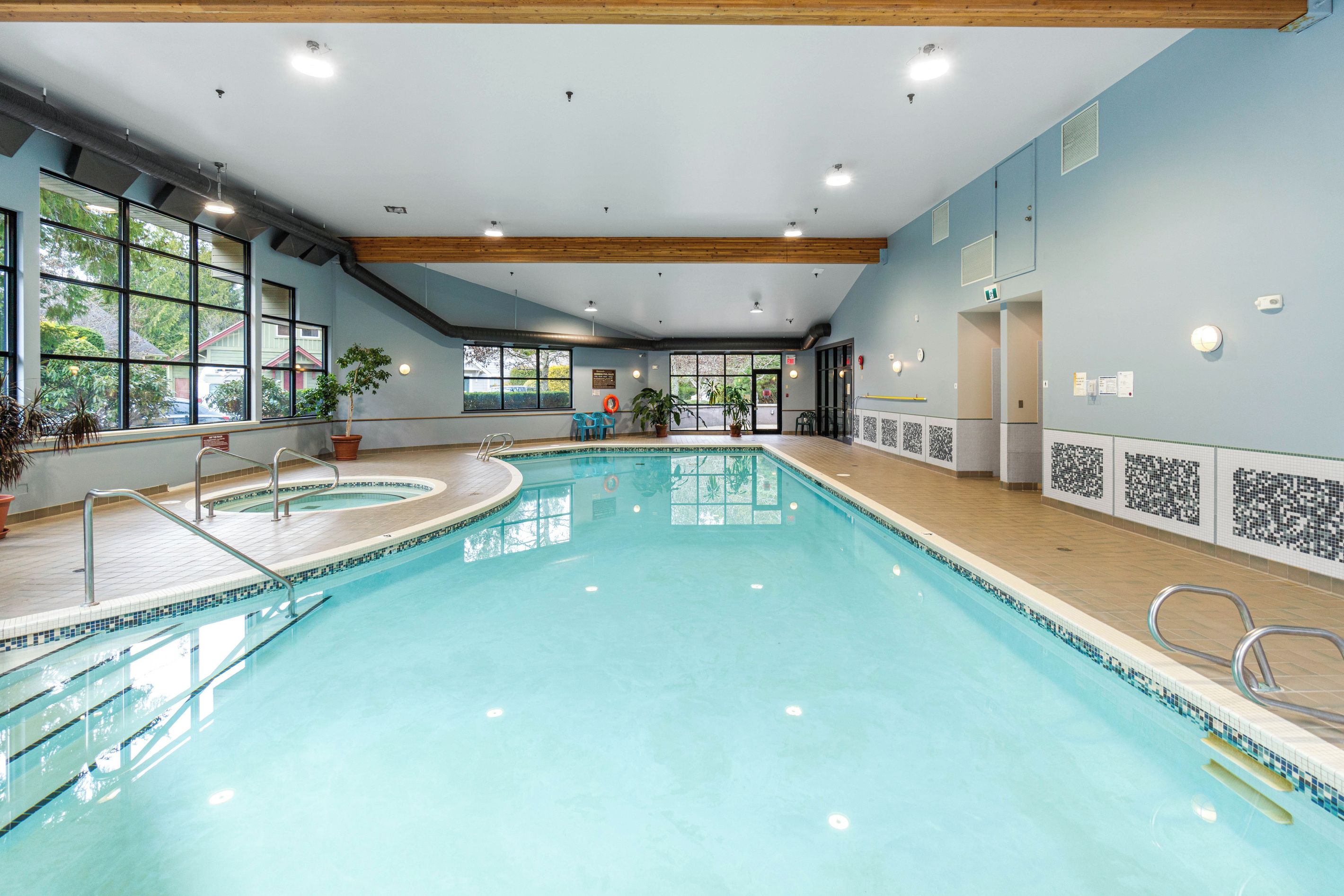 An indoor swimming pool with clear water, surrounded by windows, and equipped with safety railings.