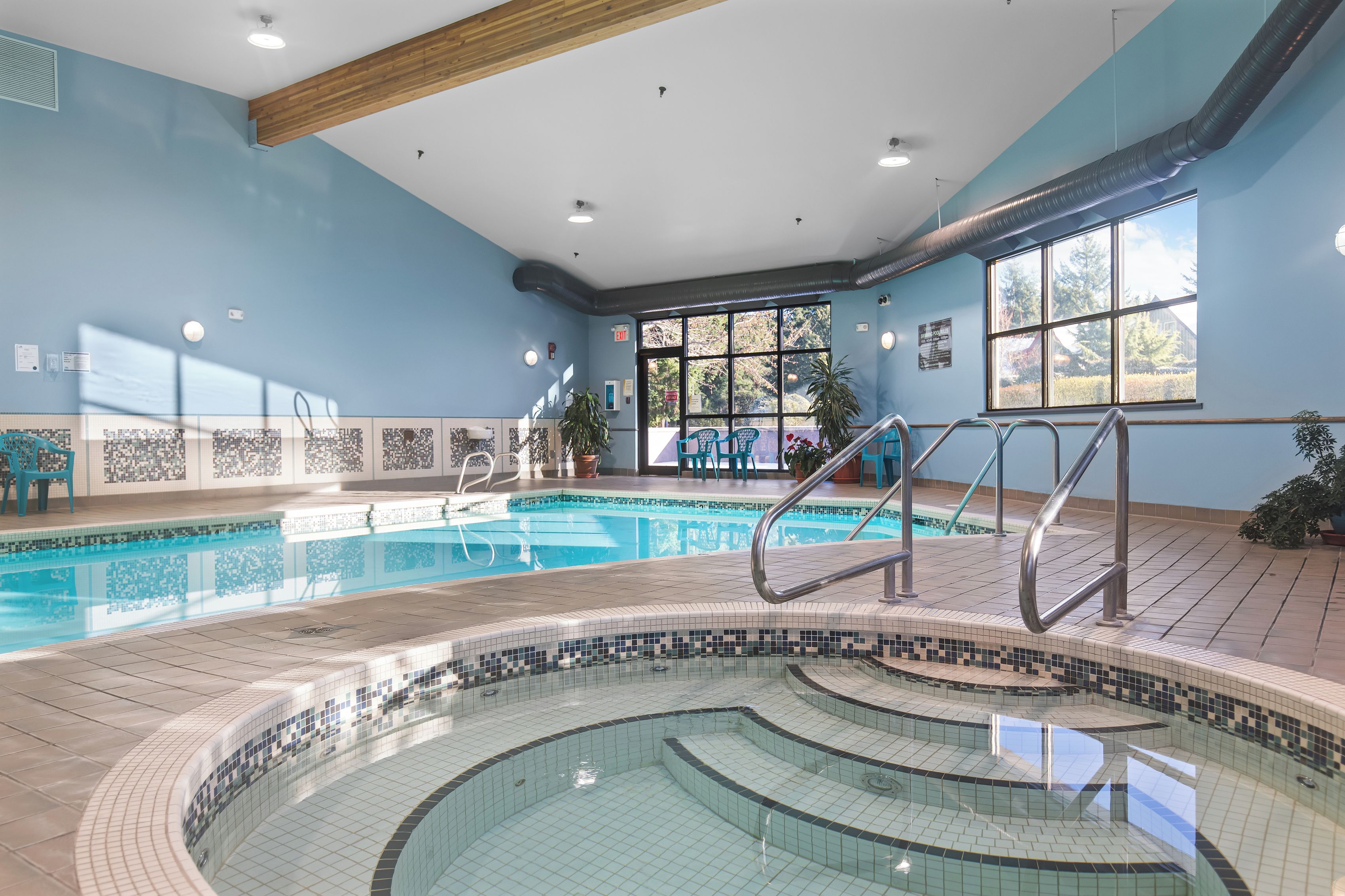 An indoor swimming pool with a tiled deck, a sunlit room with blue walls, and a view of the outdoors through large windows.