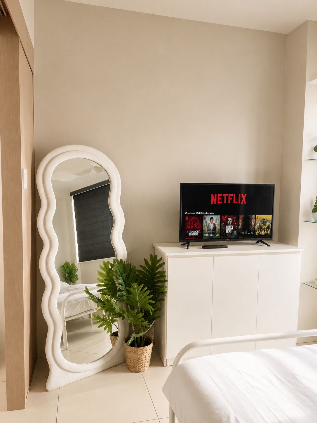 A hotel room with a bed, a white cabinet with a television displaying Netflix, a large mirror, and a potted plant.