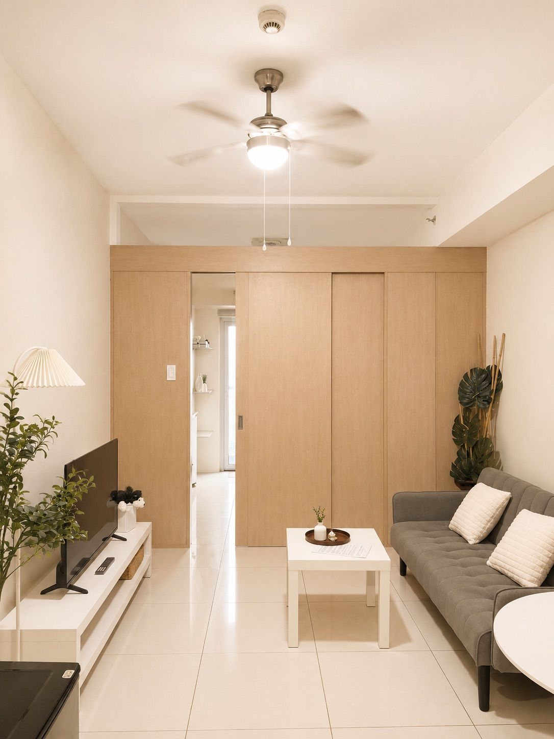 A modern living room with a ceiling fan, a flat-screen TV on a white stand, a grey sofa with cushions, and a white coffee table.