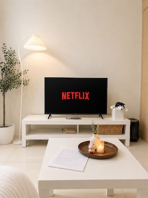 Una sala de estar moderna con un televisor de pantalla plana mostrando el logotipo de Netflix, una mesa de centro blanca con una vela y un objeto decorativo, una lámpara de pie y una planta en maceta.