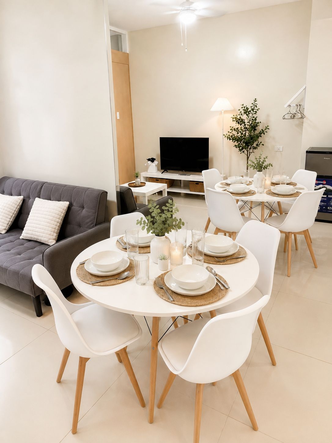 A modern living room with a grey sofa, a dining table set with white plates and glasses, and a flat-screen TV mounted on the wall.