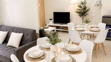 A modern living room with a grey sofa, a dining table set with white plates and glasses, and a flat-screen TV mounted on the wall.