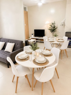 Una sala moderna con un sofá gris, una mesa de comedor con platos y vasos blancos, y un televisor de pantalla plana montado en la pared.