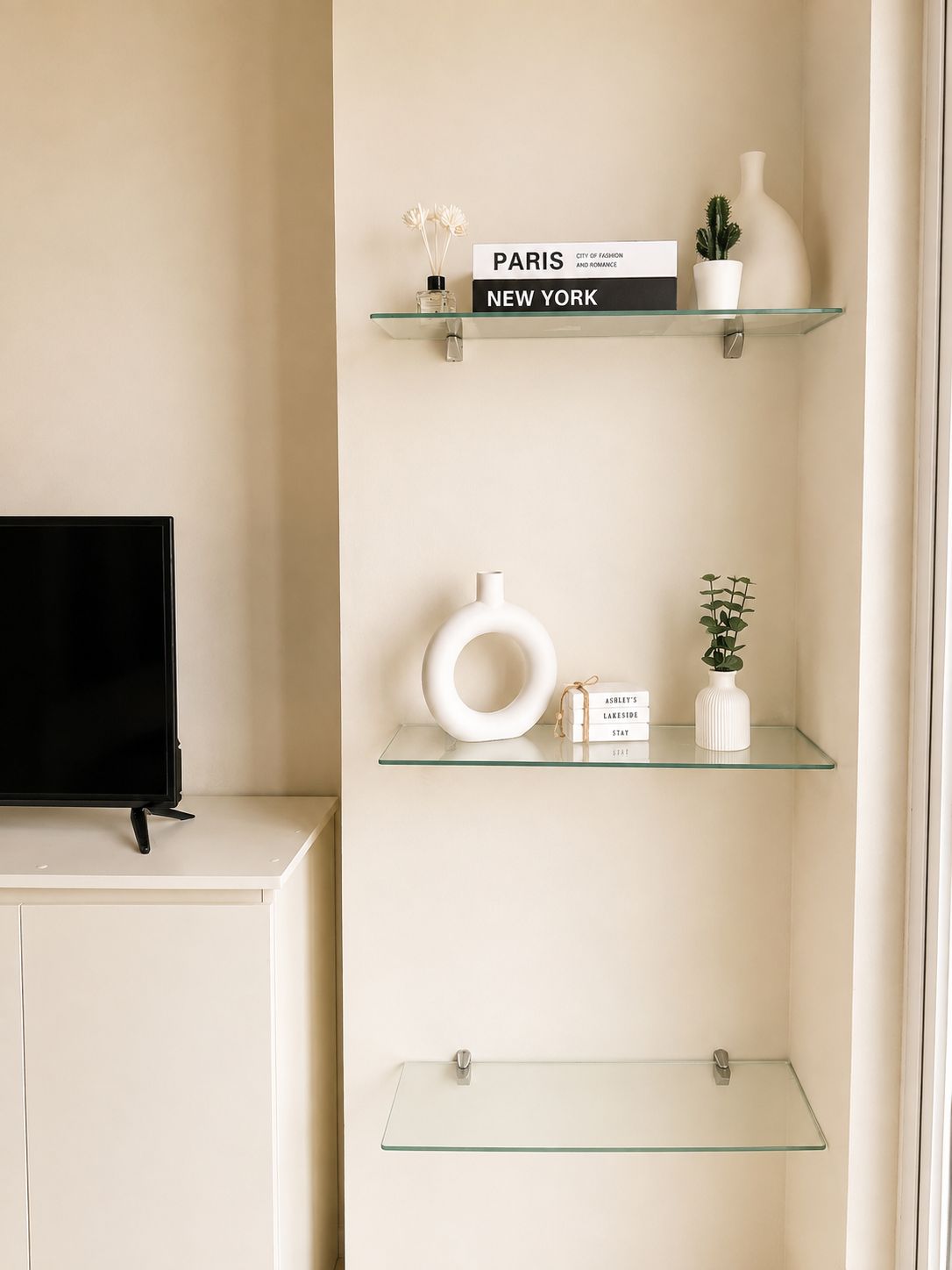 Ein schlichter Raum mit einem Flachbildfernseher auf einem weißen Schrank, drei Glasregalen mit Dekoration und einer schlichten Wand.