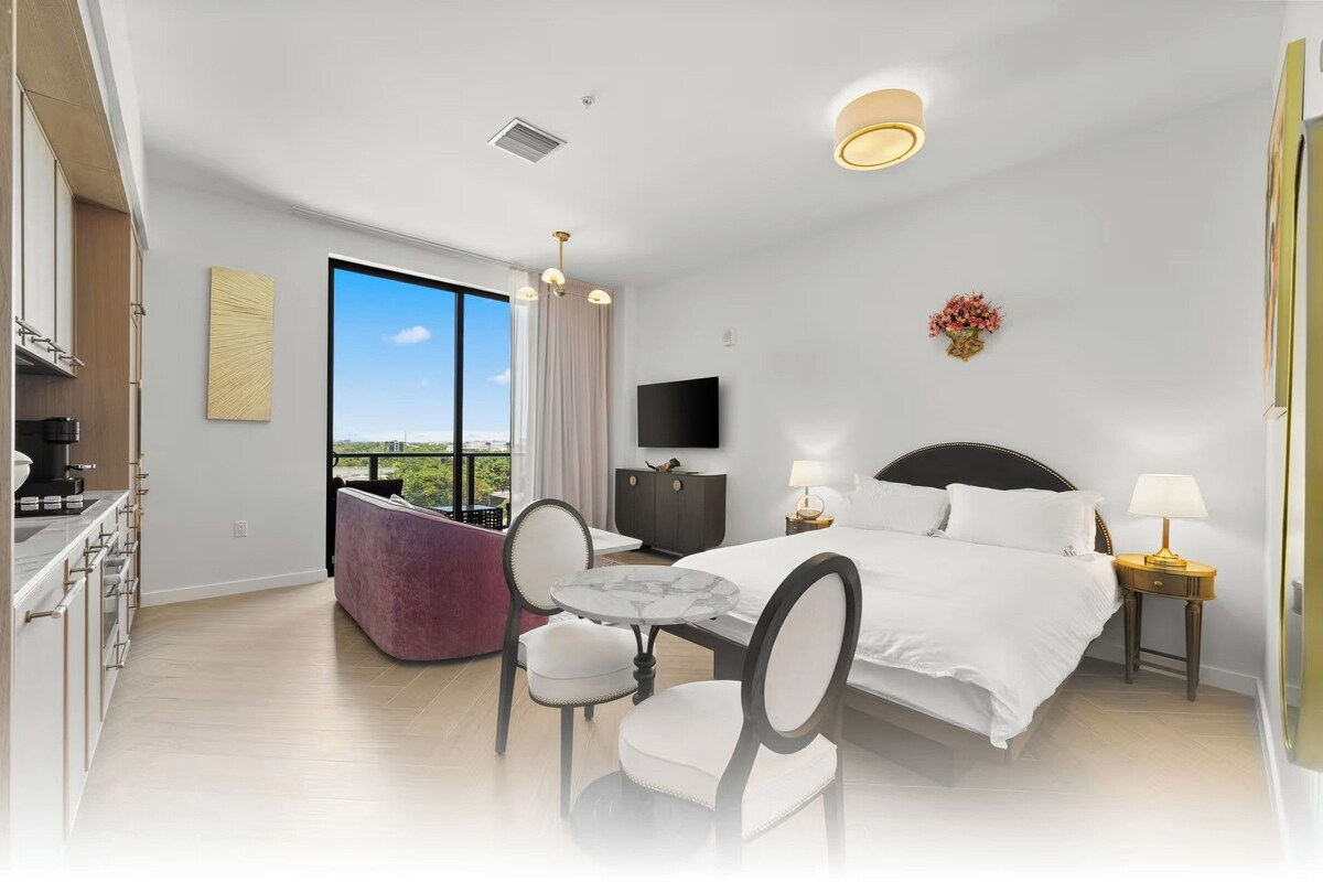 A hotel room with a bed, bedside tables, a TV, a balcony with a view, and a dining area with a table and chairs.