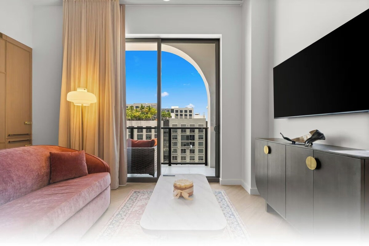 A modern hotel room with a sofa, a coffee table, a TV, and a view of buildings through a glass door.