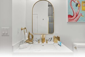 A bathroom with a large mirror, a white sink, gold fixtures, and a painting of flamingos. - NoMad Residences Wynwood Private Luxury Suites (Miami)