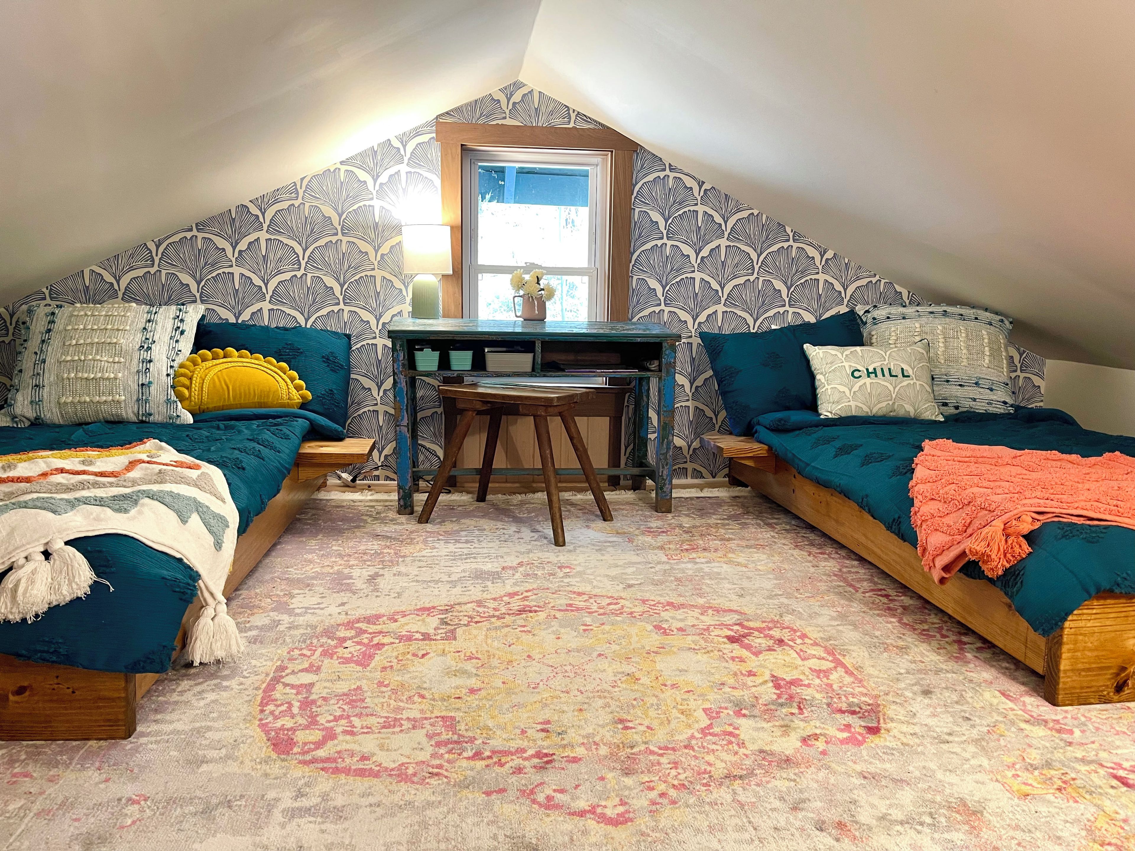 A cozy attic bedroom with a sloped ceiling, two beds with colorful bedding, a small table, and a patterned rug.