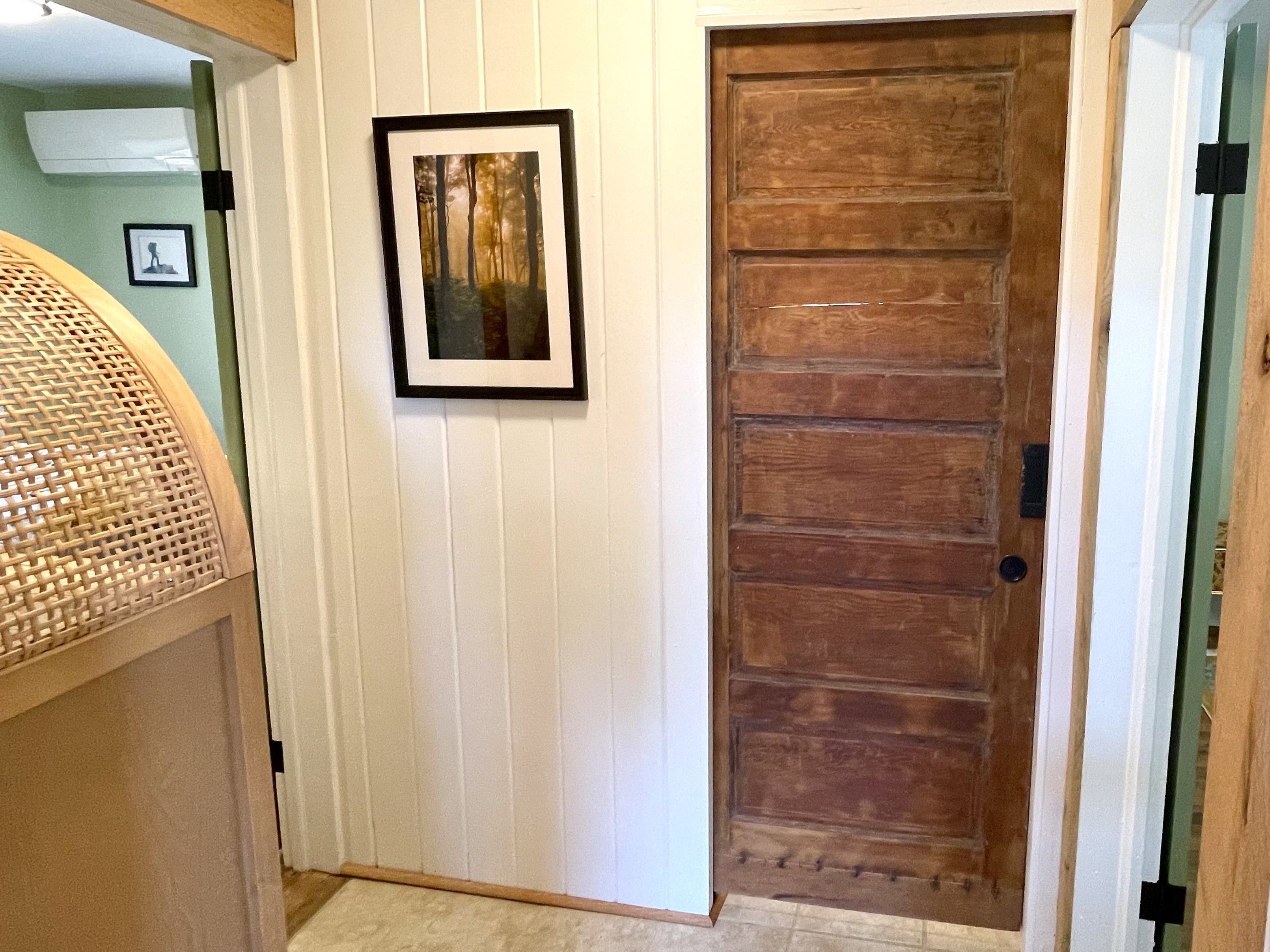 A hallway with a wooden door, a framed picture on the wall, and a wicker chair.