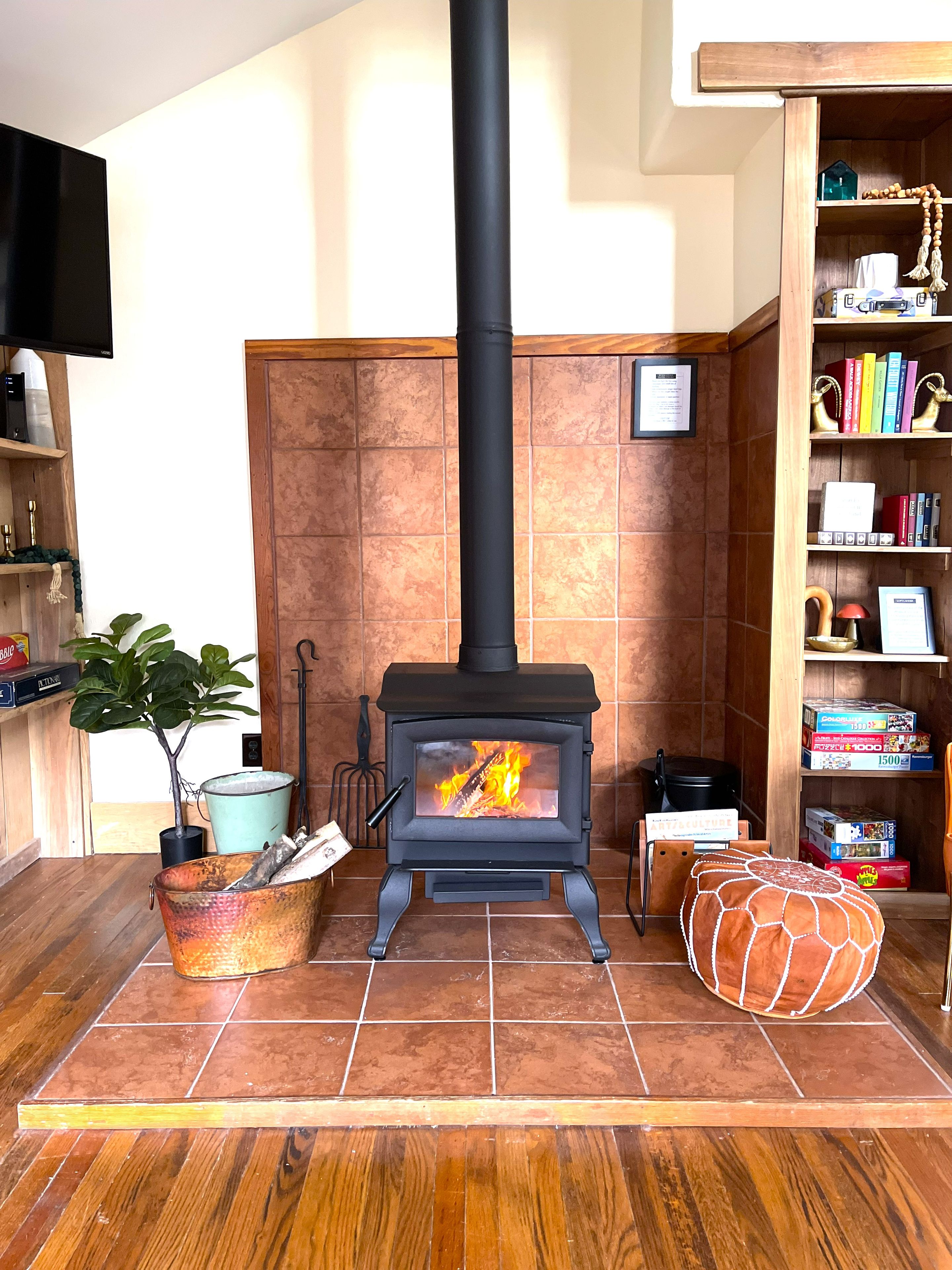 Een kamer met een houtkachel, een aan de muur bevestigde televisie en een boekenplank vol boeken en decoratieve objecten.