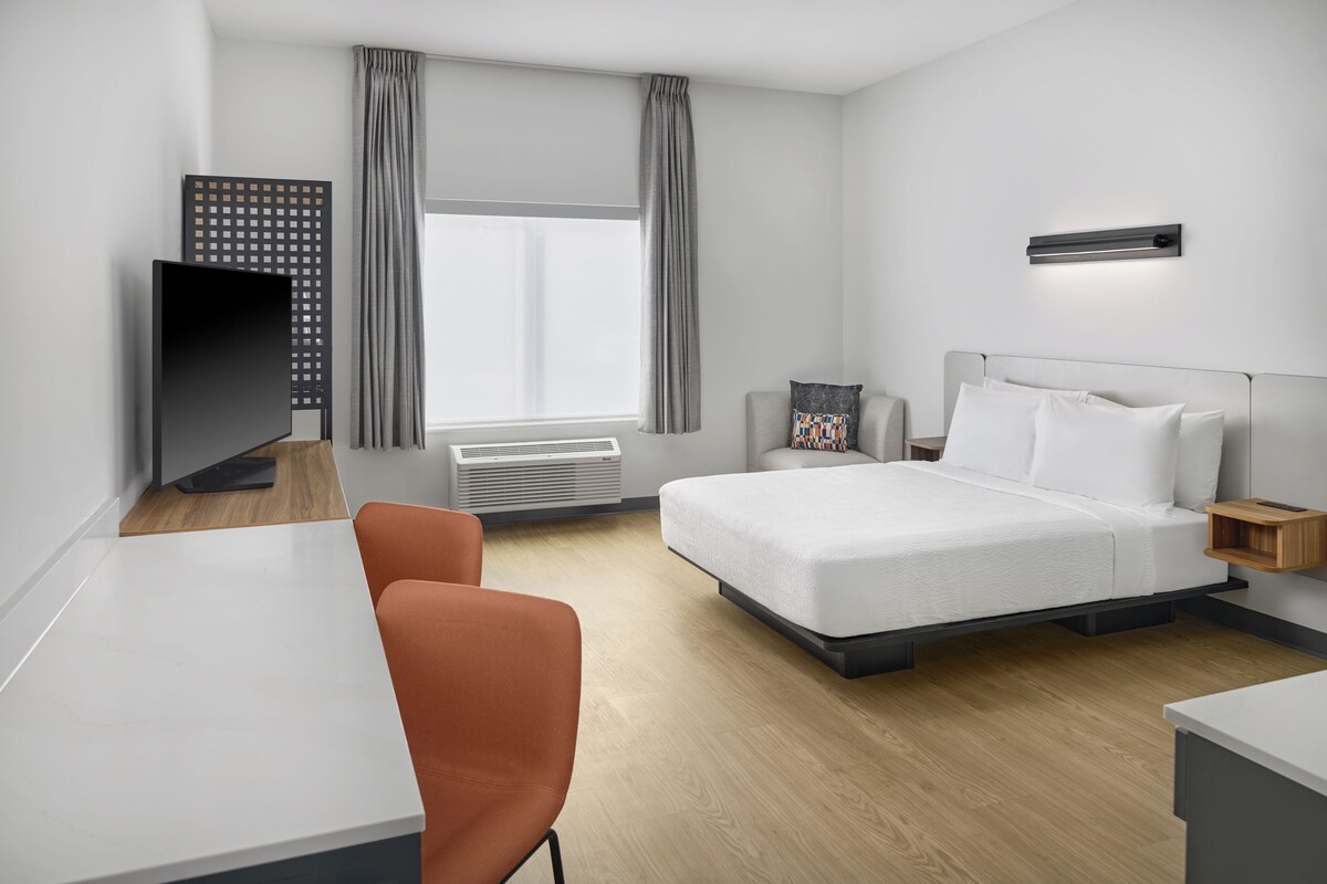 A modern hotel room with a bed, a desk, a chair, a TV, and a window with curtains.