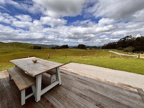 A wooden deck with a picnic table and a bowl, overlooking a grassy field with a fence and distant hills.