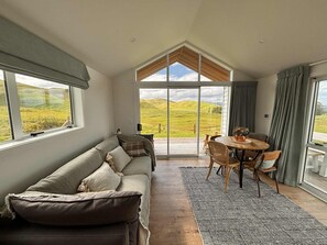A spacious room with a large window, a wooden floor, a sofa, a dining table with chairs, and a view of green hills.