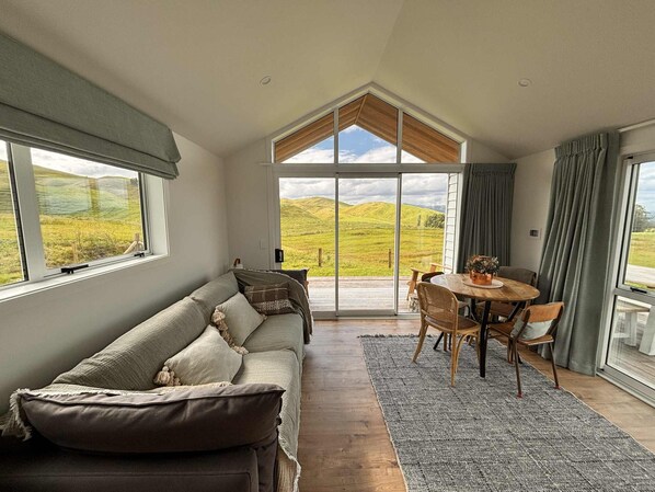A spacious room with a large window, a wooden floor, a sofa, a dining table with chairs, and a view of green hills.