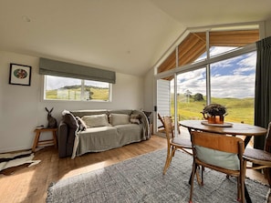 A cozy living room with a sofa, a dining table, and chairs, a large window with a view of the countryside, and a small table with a plant.