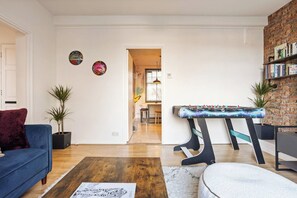 A living room with a blue sofa, a coffee table, a foosball table, and a bookshelf against a brick wall. - Design-Led Shoreditch Flat | Graffiti • Brick Lane (Greater London)