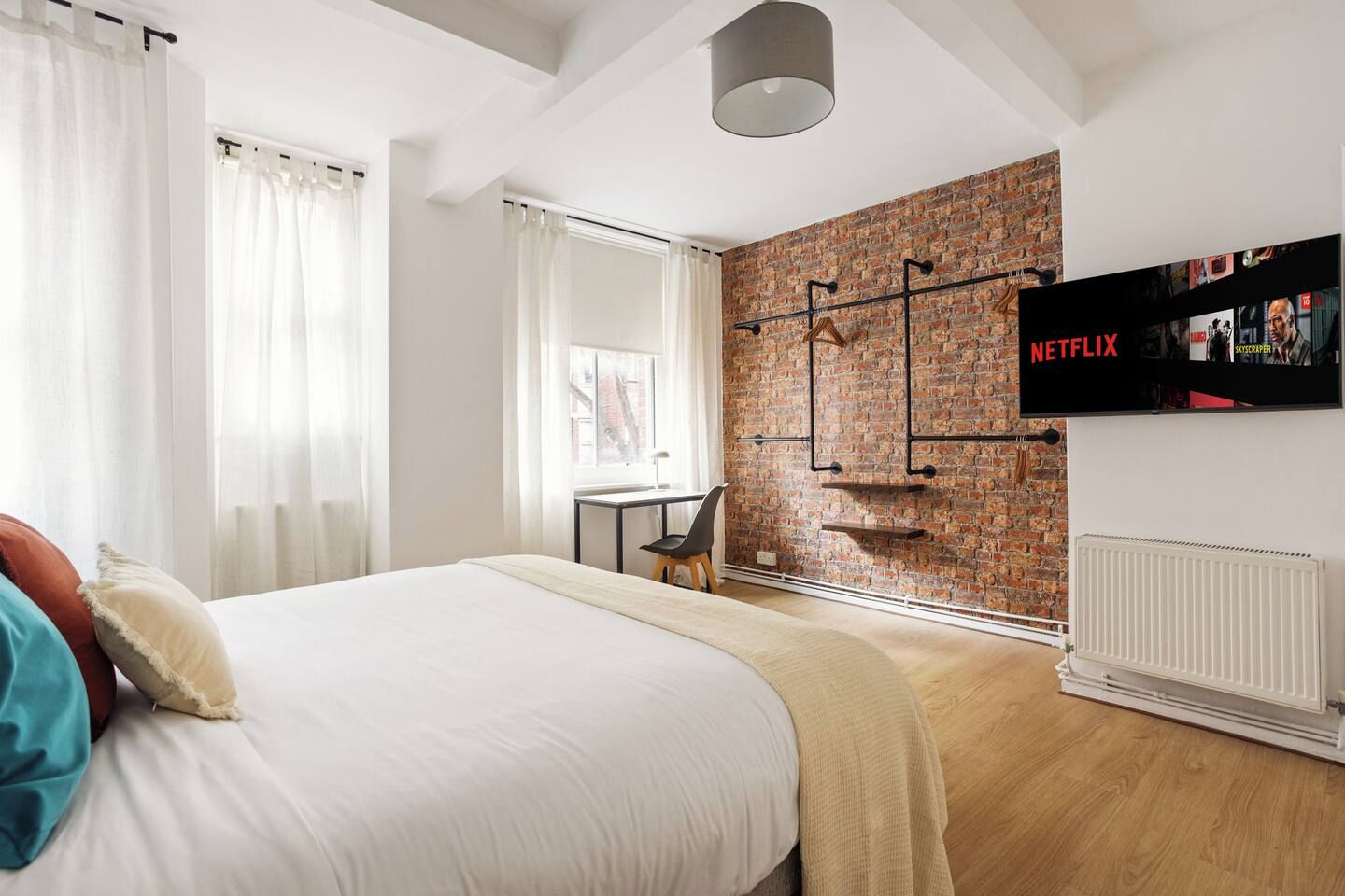 A modern hotel room with a large bed, a desk, a brick accent wall, and a mounted television displaying the Netflix logo.