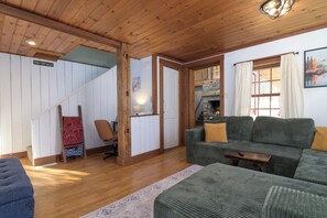 A cozy living room with a wooden ceiling, a staircase, a green sofa with cushions, and a small table.