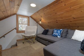 A cozy loft bedroom with a gray sofa, a white chair, and a wooden ceiling. - Historic 1880s Cape, 10 Min to Jay Peak | Rustic Charm, Clawfoot Tub & Workspace (Montgomery Center)